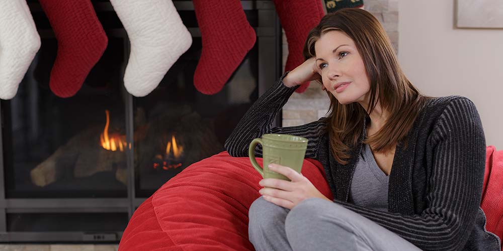 An image of a woman sitting on a Comfy Sack with a cup in her hand near a fireplace